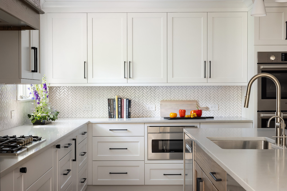 The cabinets and drawers feature minimalist pulls by Emtek. Countertops are Caesarstone London gray quartz with a waterfall edge. An under-counter Sharp microwave keeps countertops free of clutter. The herringbone patterned backsplash tile is from The Tile Lady Designs in Berkeley Heights.